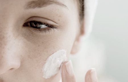 Mujer aplicándose crema en la mejilla izquierda.